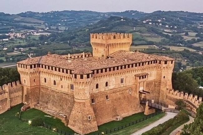 Veduta della Rocca di Gradara nelle Marche, una maestosa fortezza medievale in mattoni situata su una collina. La struttura presenta torri angolari, un mastio centrale e camminamenti di ronda merlati, circondati da una doppia cinta muraria e aree verdi curate, con le colline marchigiane sullo sfondo.