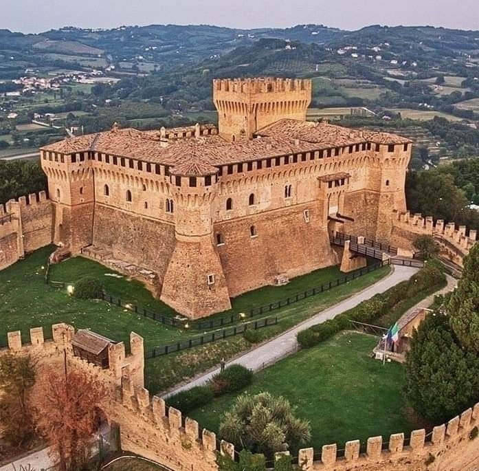 Veduta della Rocca di Gradara nelle Marche, una maestosa fortezza medievale in mattoni situata su una collina. La struttura presenta torri angolari, un mastio centrale e camminamenti di ronda merlati, circondati da una doppia cinta muraria e aree verdi curate, con le colline marchigiane sullo sfondo.